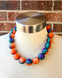 Betty chunky bead necklace with glossy orange, teal and purple acrylic beads and a silver-tone clasp on wood featured on a white mannequin neck with brick background.
